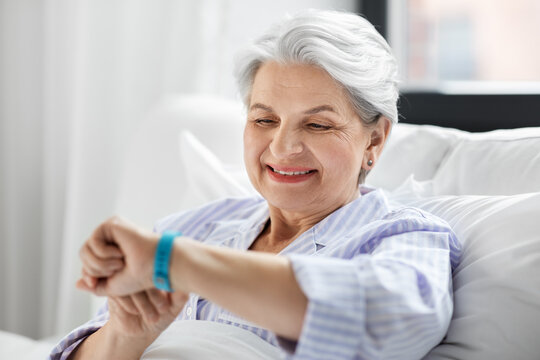 old age and people concept - happy smiling senior woman in pajamas with health tracker sitting in bed at home bedroom