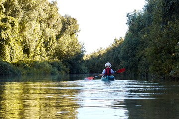 Woman row in blue kayak at wilderness areas of Danube river near green trees and thickets of wild grapes at spring