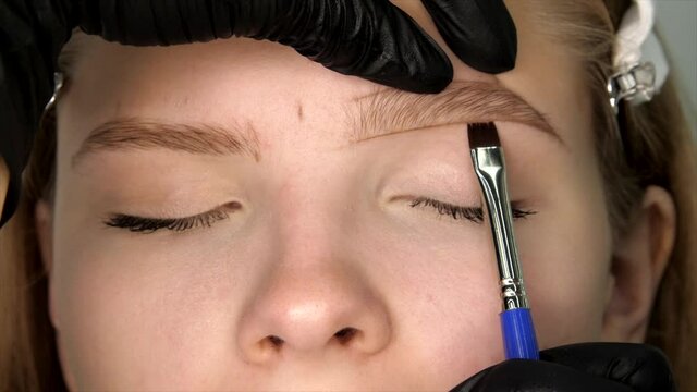 Gloved master shaping eyebrow with a cosmetic brush close-up. Master erases unnecessary sections of the line of shape of eyebrow. Shape of the eyebrows is drawn with a pencil on the female face. 
