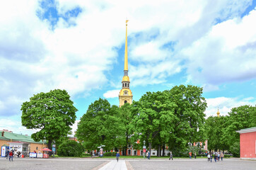 Fototapeta na wymiar The spire of the Peter and Paul Cathedral is illuminated by the sun. The cathedral is hidden in dense green trees. Bright sky with clouds. Tourists walk around the square.