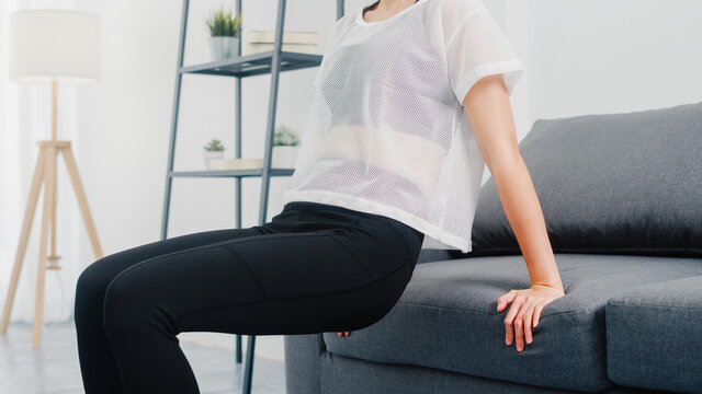 Young Korean Lady In Sportswear Exercises Doing Working Out Doing Tricep Dips Leaning On Couch In Living Room At Home. Social Distance, Isolation During The Virus. Exercises For The Lower Body.