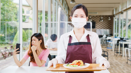 Young Asia female restaurant staff wearing protective face mask holding food tray to serving meal...