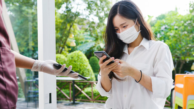 Young Asia female customer wear face mask making contactless mobile phone payment and take out food bag order from restaurant staff in coffee shop. Lifestyle new normal after corona virus concept. - Powered by Adobe