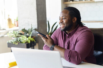 Smiling African-American man with locks hair sending voice message sitting at home office, positive black guy in casual shirt using voice recognition app, recording audio report, side view