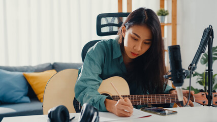 Happy asia woman songwriter play acoustic guitar listen song from smartphone think and write notes lyrics song in paper sit in living room at home studio. Music production at home concept.