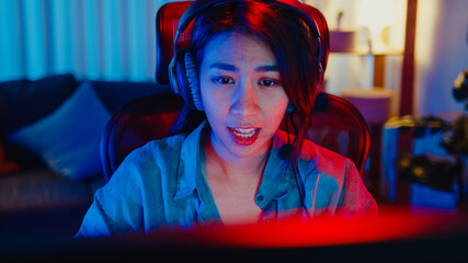 Close-up face happy asia professional girl gamer wear headphone competition play video game neon light computer in living room at home. Esport streaming game online, Home quarantine activity concept. © tirachard