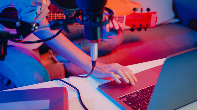 Happy asia girl blogger play guitar and use microphone sing song record music sound mixer on laptop in modern living room home studio at night. Music content creator, Tutorial, Broadcast concept.