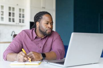 Serious and focused African-American guy watching webinars on the laptop and taking notes, studying...