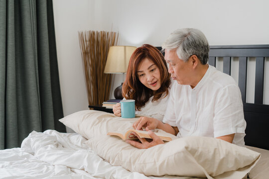 Asian senior couple talking on bed at home. Asian Senior Chinese grandparents, husband and wife happy drink coffee and read book after wake up while lying on bed in bedroom at home in morning concept. - Powered by Adobe