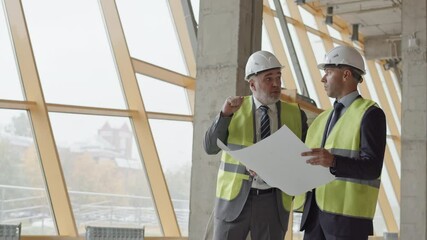 Medium long of two middle-aged male Caucasian architects wearing green reflective vests and white hard hats, standing by windows at construction site, having discussion over papers