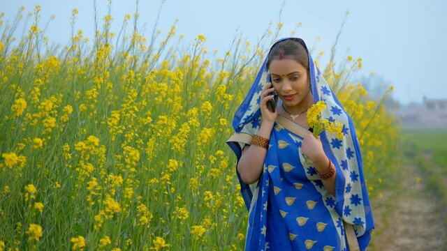 Married Indian woman talking on mobile while holding a bunch of mustard flowers. Medium shot of a young and rich village lady farmer in a saree - tech-savvy villager in mustard/Sarso fields