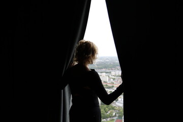 A woman standing in the dark by the window and opening the curtains with two hands