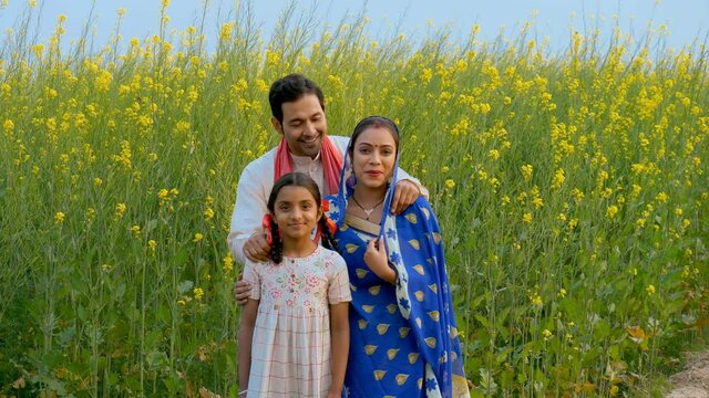Cheerful Indian village family standing in front of their mustard (Sarso) field. Medium shot of a married couple with their little daughter  smiling and posing towards the camera - village scene