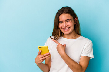 Young caucasian woman holding a mobile phone isolated on blue background smiling and pointing aside, showing something at blank space.