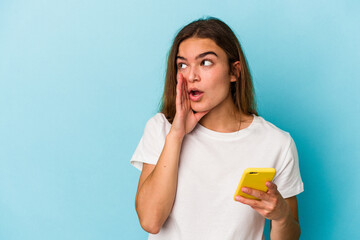 Young caucasian woman holding a mobile phone isolated on blue background is saying a secret hot braking news and looking aside