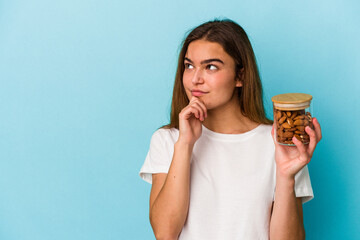 Young caucasian woman holding an almond jar isolated on blue background looking sideways with doubtful and skeptical expression.