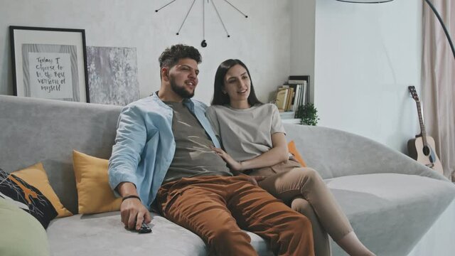 Medium PAN shot of happy young Latin couple sitting on sofa in modern living room and laughing while watching comedy on TV having movie night together at home