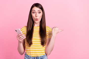 Photo of excited young teenager lady point empty space hold telephone wear orange t-shirt isolated on pastel pink color background