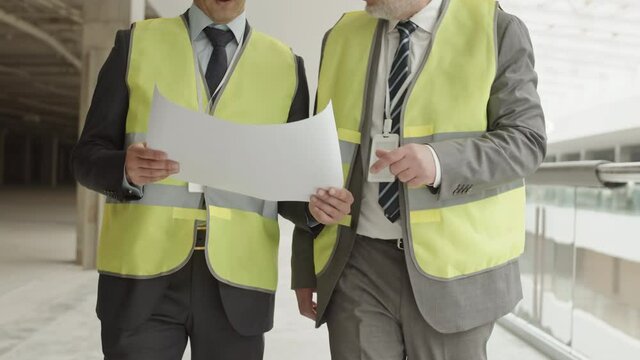 Tracking shot of two cropped unrecognizable construction workers wearing formal suits and reflective vests, holding project document, walking on site