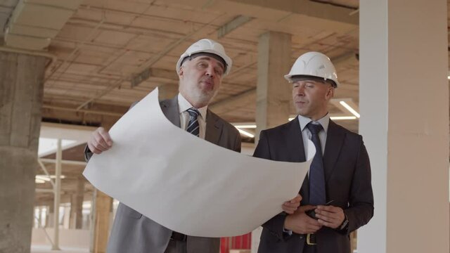 Medium tracking of two Caucasian foremen wearing formal suits and white hard hats, walking around construction site, holding project plan on paper, talking