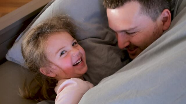 Smiling father plays with little preschooler girl lying on large bed with grey pillows and plays at home close upper view
