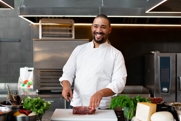Handsome young African chef standing in professional kitchen in restaurant preparing a meal of meat and cheese vegetables.