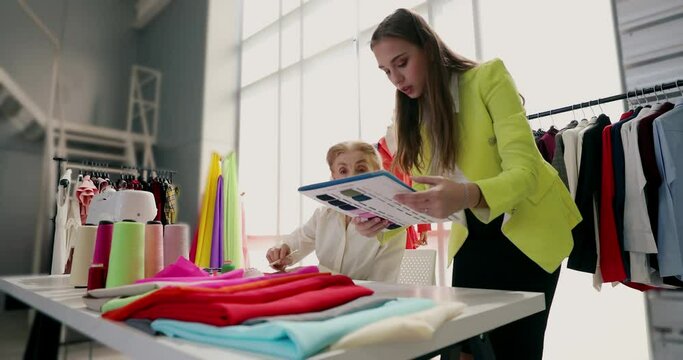 Young junior girl and senior old woman designers busy working together and discuss fashion colors in a dressmaking clothes shop. Concept of entrepreneurial and startup for small b
