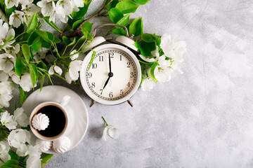  White alarm clock and a bouquet of white apple flowers and a cup of coffee on the light background, summer morning concept. Free space for text