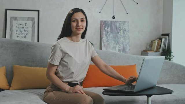 Medium PAN portrait of smiling Hispanic or Latin woman sitting on light-grey sofa in cozy living room and working on laptop then looking at camera smiling