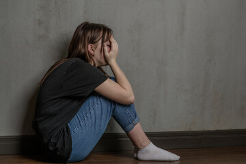Crying teen girl sits on the floor near a wall and covered her face with her hands. Empty space for...