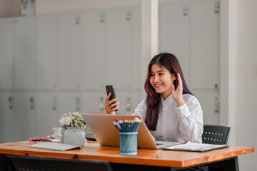 Happy young asian woman holding phone looking at screen waving hand video calling distance friend online in mobile chat app using smartphone videochat while working with laptop computer at cafe