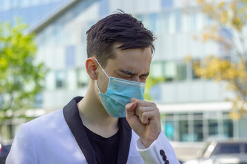 Street portrait of a young man 18-21 years old coughing and covering his mouth in a medical mask against the background of a glass office building.