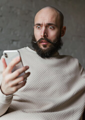 bald man 35 years old with a beard and mustache in a beige jumper looks at the phone with surprise and fear