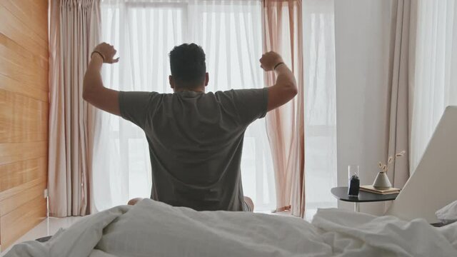 Rear-view medium shot of young man of Latin ethnicity getting up in morning and stretching on cozy bed looking at panoramic window