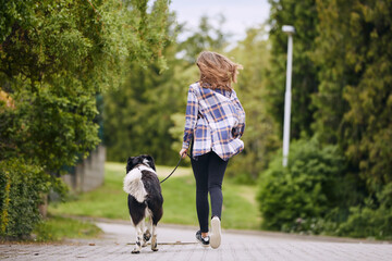 Naklejka na ściany i meble Rear view of teenage girl running with her dog on pet leash down street..