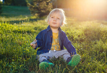 Portrait of a cute toddler boy sitting in a field among the grass at sunset. A child walks in the park. On open air. Happy summer and lifestyle concept. Childhood.