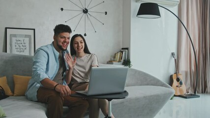Medium shot of cheerful young couple sitting on sofa in front of laptop having video chat with friends or family - Powered by Adobe