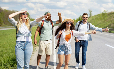 Obraz premium Happy multiethnic friends trying to get free ride, hitchhiking on highway, showing thumb up gestures outdoors, panorama
