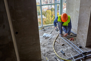 Worker or builder in workwear and safety helmet is installing plastic pipes using modern tools in a flat of building under construction