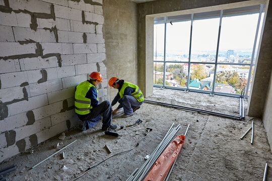 Two Men Builders In Workwear And Hardhats Installing Plastic Pipes Using Modern Tools In Flat Of Building Under Construction With Panoramic Cityscape