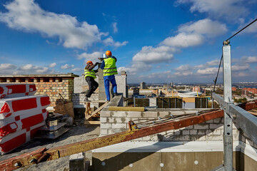 Two workers in protective clothing and safety helmets are working with ventilation system on roof of building under construction - obrazy, fototapety, plakaty