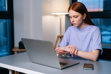 Overworked redhead young woman typing on laptop actively feeling pain in wrist sitting at desk near window in evening. Lady working on laptop and suffering from pain in hand, carpal tunnel syndrome.