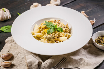 Close-up penne pasta with mushrooms, parmesan cheese sauce and fresh basil in a large white plate. Italian food still life on a dark table. Traditional Mediterranean cuisine.