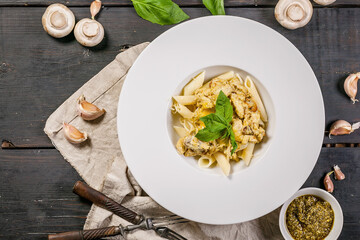 Penne pasta with mushrooms, parmesan cheese sauce and fresh basil in a large white plate. Italian food still life on a dark table. Traditional Mediterranean cuisine. Top view