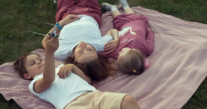 Three members of one family lie on a blanket on the grass. This is a mother and her two children. The woman and her daughter look at each other, then turn to the boy who is playing an airplane model