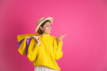 Beautiful woman in bright yellow sweater and straw hat on pink background hold shopping bags happy...