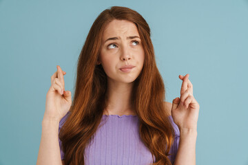 Young ginger woman frowning while holding fingers crossed for god luck