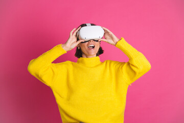 Beautiful woman in bright yellow sweater on pink background in virtual reality glasses happy excited overjoyed touch air