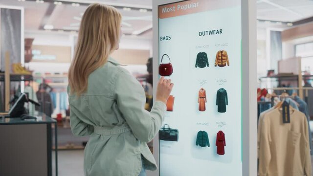 Beautiful Female Customer Using Floor-Standing LCD Touch Display while Shopping in Clothing Store. She is Choosing Stylish Bags, Picking Different Designs from Collection. People in Fashionable Shop.