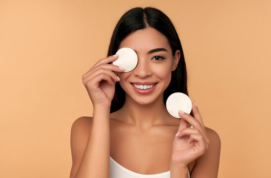 Young woman cleans beauty face using cotton pads, cleansing lotion and face toner to remove makeup. Nice girl with beautiful skin. Skin care concept. Beige background, copy space